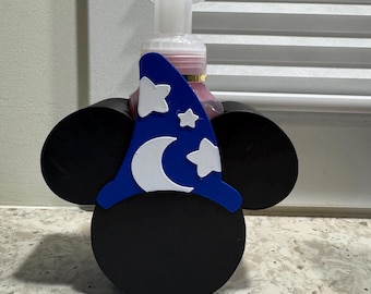 May include: A black wooden soap dispenser holder shaped like Mickey Mouse ears, with a blue wizard hat featuring a white crescent moon and stars. The dispenser has a white pump and a pink liquid inside.