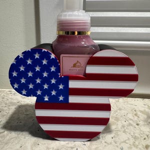 May include: A decorative soap dispenser holder in the shape of Mickey Mouse ears, featuring the American flag design. The ears are red and white striped, and blue with white stars. A pink soap dispenser is visible behind the holder.