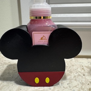 May include: A black and red Mickey Mouse-shaped soap dispenser holder. The holder has two yellow circles on the red bottom. A pink soap bottle with a gold rim is inside the holder. The bottle has a white label with text.