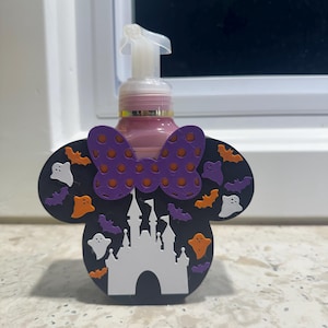 May include: A Halloween-themed soap dispenser featuring a black Mickey Mouse head shape with a purple polka-dot bow. The design includes orange ghosts, bats, and a white castle. The dispenser has a pink liquid inside.
