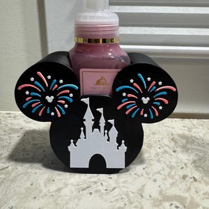 May include: A black soap dispenser holder shaped like Mickey Mouse ears, with a white castle silhouette and colorful fireworks accents. The holder cradles a pink soap bottle with a white pump.