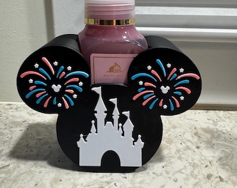 May include: A black soap dispenser holder shaped like Mickey Mouse ears, with a white castle silhouette and colorful fireworks accents. The holder cradles a pink soap bottle with a white pump.