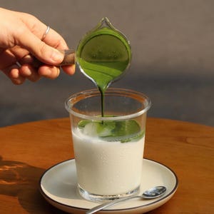 May include: A clear plastic cup filled with a white liquid, possibly milk, sits on a white plate with a silver spoon. A small glass pitcher with a wooden handle pours bright green liquid into the cup. The drink is on a wooden table.