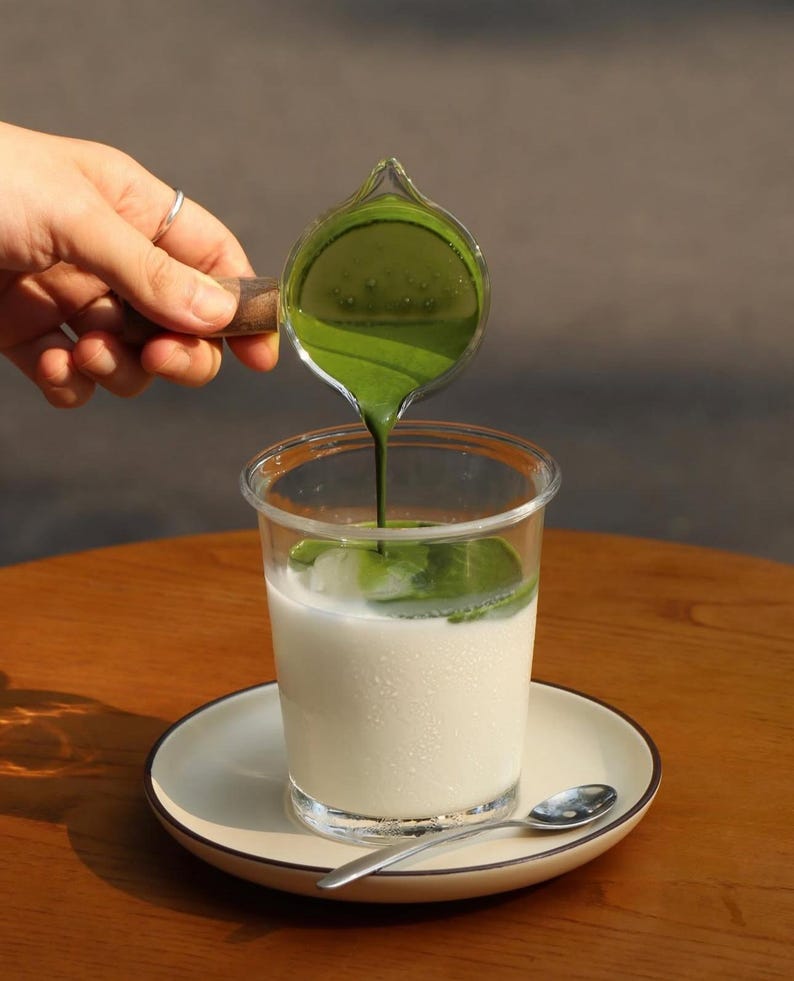 May include: A clear plastic cup filled with a white liquid, possibly milk, sits on a white plate with a silver spoon. A small glass pitcher with a wooden handle pours bright green liquid into the cup. The drink is on a wooden table.