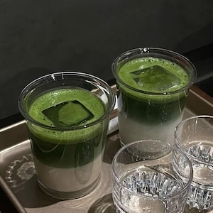 May include: Two clear plastic cups filled with layered matcha latte, topped with a square ice cube. Two additional clear glasses are in front of the drinks. The drinks are on a metal tray.