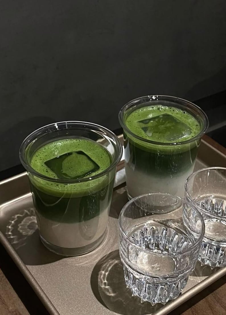 May include: Two clear plastic cups filled with layered matcha latte, topped with a square ice cube. Two additional clear glasses are in front of the drinks. The drinks are on a metal tray.