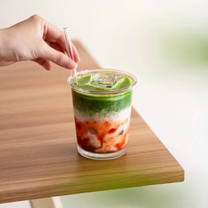 May include: A layered drink in a clear plastic cup with a clear straw. The drink has layers of green, white, and red, with ice cubes on top. The drink is on a wooden table.