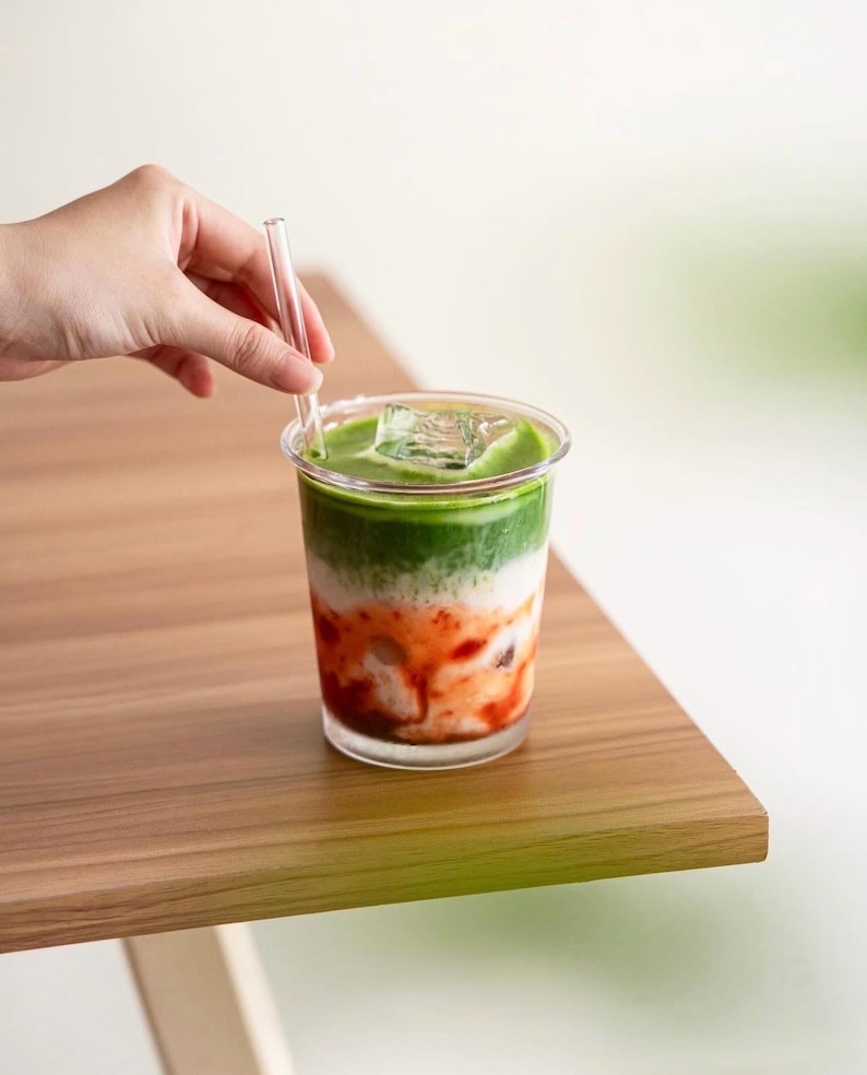 May include: A layered drink in a clear plastic cup with a clear straw. The drink has layers of green, white, and red, with ice cubes on top. The drink is on a wooden table.
