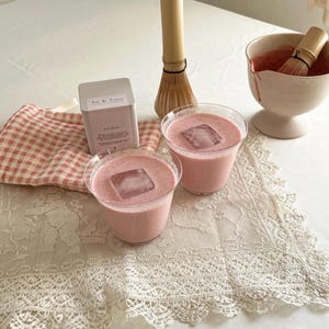 May include: Two clear glass cups filled with a pink beverage and a single square ice cube, a square metal tin labeled The de Young, a bamboo whisk, and a ceramic bowl containing pink liquid, all arranged on a white lace-trimmed fabric surface.