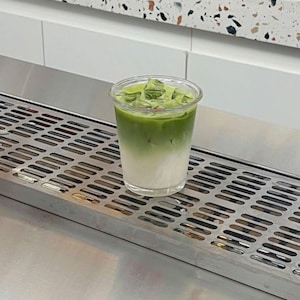 May include: A layered matcha latte in a clear plastic cup, with a gradient of white at the bottom and vibrant green at the top, filled with ice cubes. The drink sits on a stainless steel surface, with a white and speckled countertop in the background.