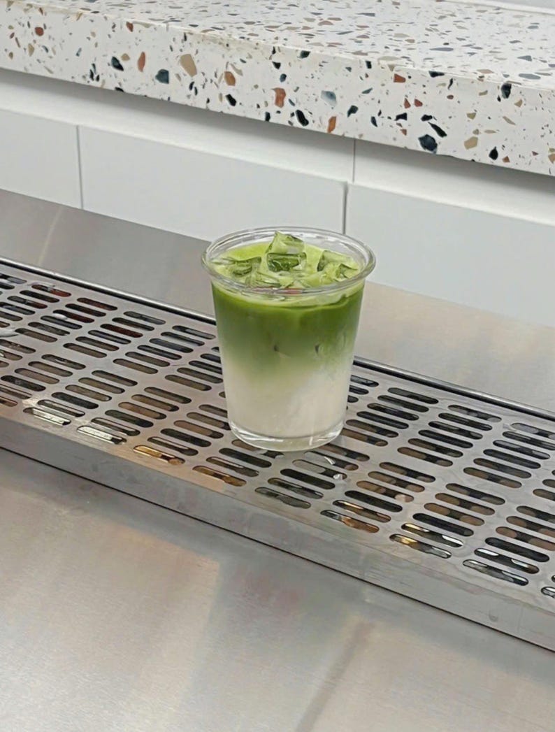May include: A layered matcha latte in a clear plastic cup, with a gradient of white at the bottom and vibrant green at the top, filled with ice cubes. The drink sits on a stainless steel surface, with a white and speckled countertop in the background.