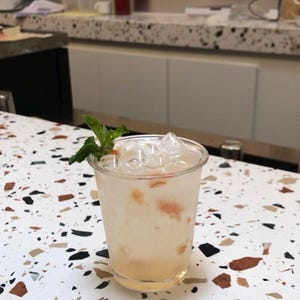 May include: A refreshing cocktail in a clear plastic cup, filled with ice cubes and a light-colored liquid, garnished with a sprig of mint. The drink sits on a speckled countertop with various colors.
