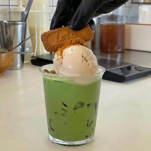 May include: A clear plastic cup filled with a vibrant green beverage, topped with a scoop of light brown ice cream and cookie crumbs. A hand in a black glove is placing a cookie on top. The cookie has the word "Lotus" on it.