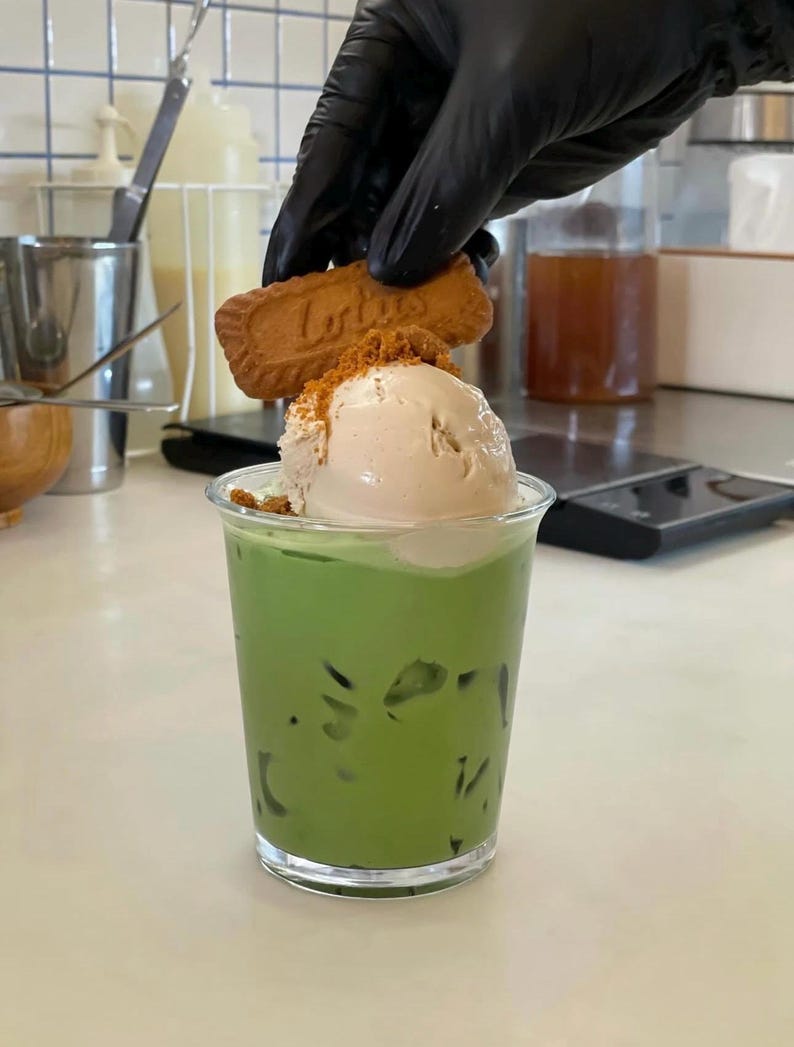 May include: A clear plastic cup filled with a vibrant green beverage, topped with a scoop of light brown ice cream and cookie crumbs. A hand in a black glove is placing a cookie on top. The cookie has the word "Lotus" on it.
