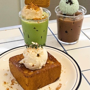 May include: An overhead shot of three desserts. A slice of golden-brown toast topped with ice cream and nuts sits on a plate. Two clear cups hold drinks, one green with ice cream and a cookie, the other brown with mint ice cream.