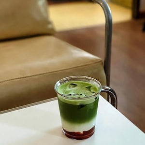 May include: A layered beverage in a clear glass with a handle. The drink features a red base, a white middle layer, and a green top layer with ice cubes. The glass sits on a white table, with a blurred background.