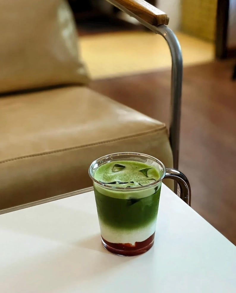 May include: A layered beverage in a clear glass with a handle. The drink features a red base, a white middle layer, and a green top layer with ice cubes. The glass sits on a white table, with a blurred background.