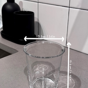 May include: A clear glass tumbler, 3.6 inches wide and 4.09 inches tall, sits on a gray countertop. A black soap dispenser and a black cup are in the background. The tumbler is empty and transparent.