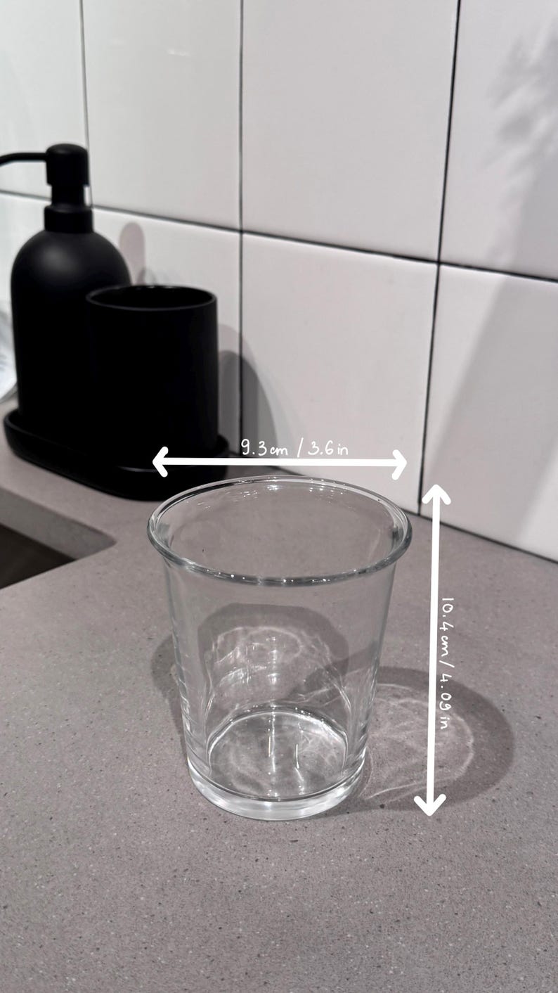 May include: A clear glass tumbler, 3.6 inches wide and 4.09 inches tall, sits on a gray countertop. A black soap dispenser and a black cup are in the background. The tumbler is empty and transparent.
