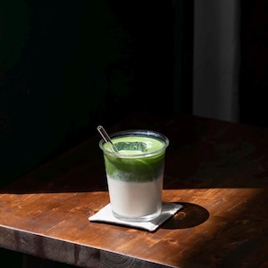 May include: A layered matcha latte in a clear plastic cup with a clear straw. The drink has a white base, a green middle layer, and a green top layer. The cup sits on a small white square coaster on a wooden table.