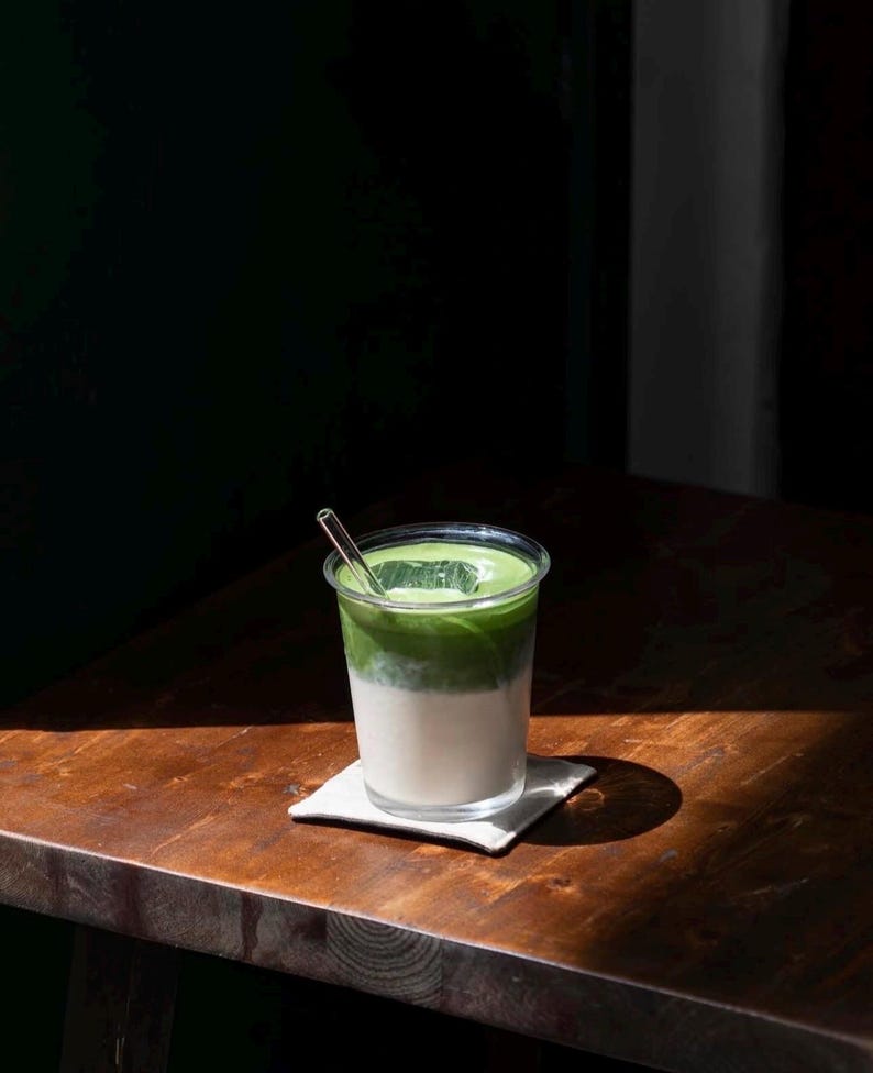 May include: A layered matcha latte in a clear plastic cup with a clear straw. The drink has a white base, a green middle layer, and a green top layer. The cup sits on a small white square coaster on a wooden table.