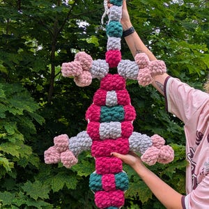 May include: A large, plush, handmade crochet lizard toy featuring a segmented body and limbs. The creature is constructed from soft, chunky yarn in alternating blocks of vibrant magenta, deep forest green, light gray, and pale pink colors.