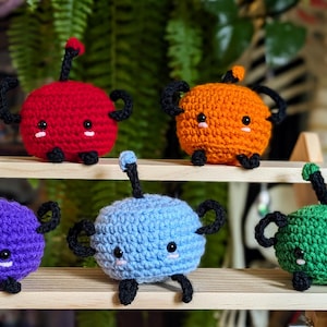 Può includere: Sei creature rotonde all'uncinetto in vari colori: rosso, arancione, viola, blu e verde. Ognuna ha braccia, gambe e uno stelo neri. Sono esposte su una mensola di legno. Misure non disponibili.