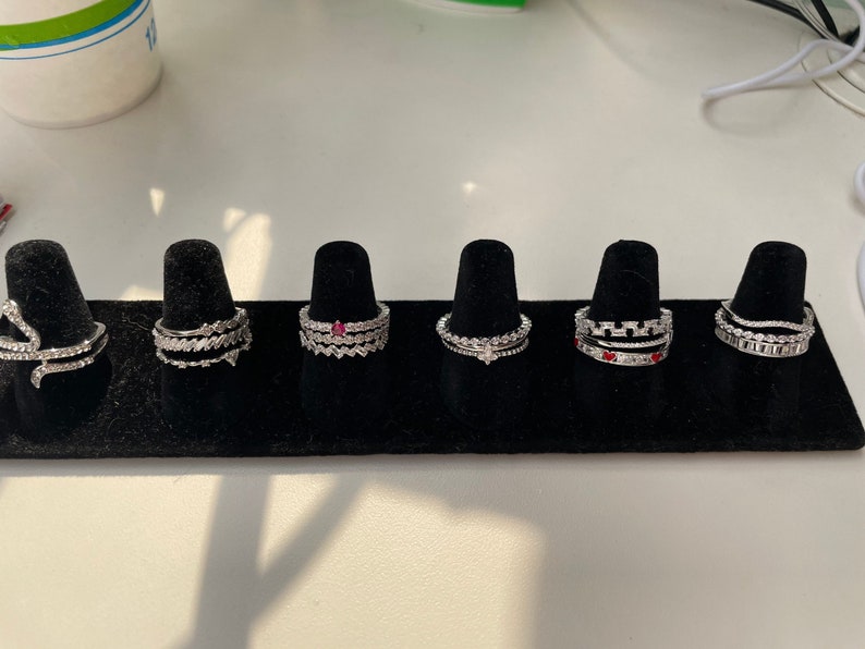 Puede incluir: Seis anillos de plata con piedras transparentes y rojas, expuestos sobre terciopelo negro. Los anillos est&aacute;n apilados uno encima del otro.