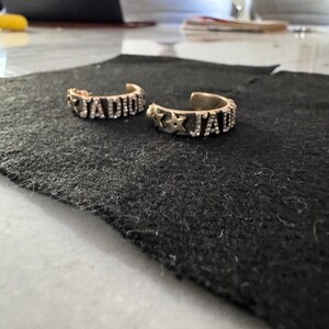 May include: Two gold-toned rings with the word "J'ADIOR" spelled out in rhinestones. The rings are on a black surface.