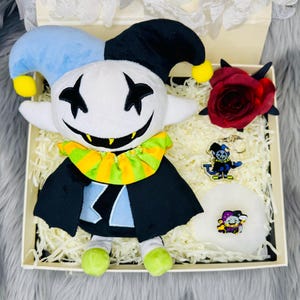 May include: A gift box containing a plush doll, a red rose, a keychain, and a white puff. The plush doll has a white face with a black smile and eyes, a blue and black jester hat, and a black cape. The keychain and puff feature a character design.