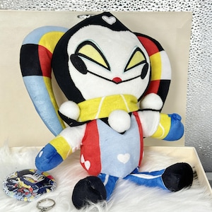 May include: A plush doll with a black and white face, yellow eyes, and a red nose. The doll wears a yellow scarf, a red and blue outfit, and has colorful ears. A button and keychain with the same character are also present.