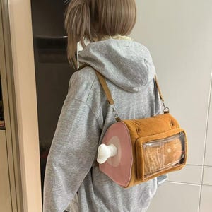 May include: A novelty shoulder bag shaped like a ham. The bag is brown with a pink and white ham design on one side and a clear window on the other. It has a brown strap and a white bone detail.