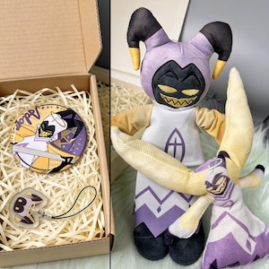 以下が含まれることがあります： 丸いピンと黒いコードのチャームが入った箱。ピンには「Adon」の文字と、黄色、紫、白のキャラクターイラストが描かれています。紫色の頭、黒い角、白と紫の衣装のぬいぐるみも写っています。