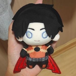 Puede incluir: Una muñeca de peluche con cabello negro y ojos azules. La muñeca lleva un atuendo naranja y negro con un cinturón gris y una capa roja. La muñeca está sujeta en la mano de una persona.