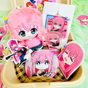 Puede incluir: Una colección de artículos de anime de color rosa, que incluye un peluche, colgantes acrílicos, pegatinas y un botón. Los artículos presentan un personaje con pelo rosa y una guitarra, presentados en una caja decorativa. La caja tiene un patrón de cuadros.