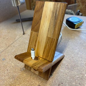 May include: A wooden phone stand constructed from light and dark wood panels with a vertical grain. The stand features a central circular cutout at the base, through which a white charging cable with a lightning connector is threaded and upright.
