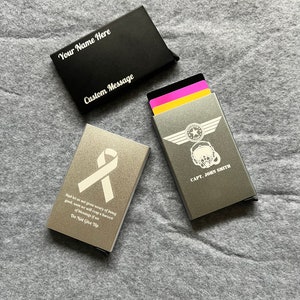 May include: Two silver metal card holders with engraved designs. One card holder has a white ribbon and the text "And let us not grow weary of doing good, soon we will reap a harvest of blessings if we Do Not Give Up". The other card holder has a pilot helmet and wings design with the text "CAPT. JOHN SMITH".