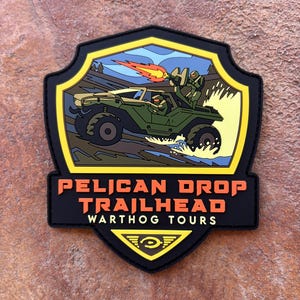 May include: A black and yellow patch with the text "PELICAN DROP TRAILHEAD WARTHOG TOURS". The patch features a green vehicle with two figures and a fiery explosion. The patch is shield-shaped.