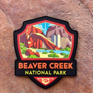 Peut inclure: Un écusson noir avec l'inscription "BEAVER CREEK NATIONAL PARK" en orange. L'écusson, en forme de bouclier, présente un paysage coloré de montagnes, d'eau et de ciel. Une bordure rouge encadre l'image.
