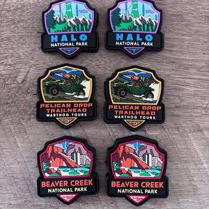 Puede incluir: Seis parches de goma en forma de escudo con ilustraciones coloridas. Dos muestran el Halo National Park con montañas, dos el Pelican Drop Trailhead Warthog Tours con un vehículo verde y dos el Beaver Creek National Park con arcos de roca rojos.