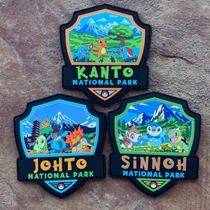 Könnte beinhalten: Drei schildförmige Aufnäher mit farbenfrohen Illustrationen von Pokémon in Nationalparks. Der obere Aufnäher zeigt KANTO NATIONAL PARK, unten links steht JOHTO NATIONAL PARK und unten rechts ist SINNOH NATIONAL PARK zu lesen.