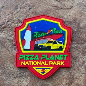 Puede incluir: Un parche rojo, amarillo y azul de Pizza Planet National Park. El parche presenta un cohete, un camión amarillo y el logotipo de Pizza Planet. El texto "Pizza Planet National Park" es verde.
