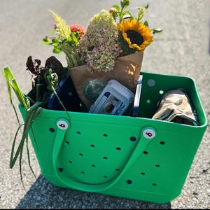 May include: A green reusable tote bag with black handles and white dots. The bag is filled with groceries, including a bouquet of flowers, leafy greens, and a brown paper bag.