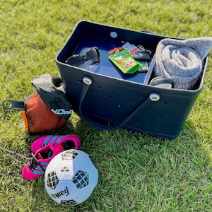 May include: A navy blue tote bag with a white polka dot pattern and a white handle. The bag is filled with various items, including a gray towel, sunglasses, a small green pouch, and a key chain. The bag is sitting on a green grassy surface.