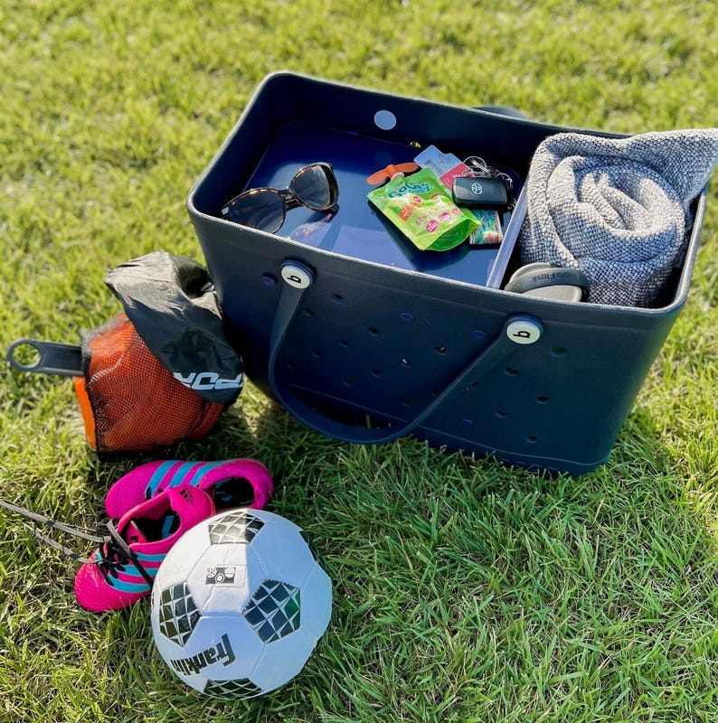 May include: A navy blue tote bag with a white polka dot pattern and a white handle. The bag is filled with various items, including a gray towel, sunglasses, a small green pouch, and a key chain. The bag is sitting on a green grassy surface.