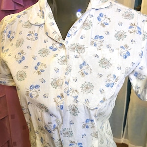 May include: White button-up shirt with a floral pattern. The shirt features short sleeves, a collared neckline, and a single chest pocket. The floral design includes blue and gray flowers.