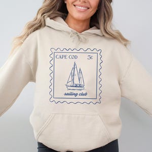 May include: A beige hooded sweatshirt featuring a blue postage stamp design. The stamp reads "CAPE COD" and "sailing club" with a sailboat illustration. The sweatshirt has a front pocket and a drawstring hood.