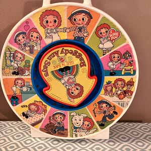 Könnte beinhalten: Ein vintage Spielzeugrad aus Plastik mit einem rot-weiß-blauen Design. Das Rad zeigt ein Bild einer rothaarigen Puppe in verschiedenen Szenen, darunter das Spielen mit einem Hund, das Schlafen und das Teetrinken. Das Rad dreht sich und hat einen kleinen Griff oben. Der Text "See 'n Say" und "Raggedy Ann" ist auf dem Rad aufgedruckt.