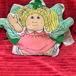 May include: A Cabbage Patch Kid plush pillow with a green cabbage leaf design. The doll is wearing a pink dress and has blonde hair with pigtails.