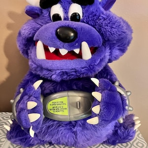 May include: Purple plush monster toy with a large mouth and sharp teeth. The monster is holding a silver object with a button that says "Push button to open handcuffs. Appuyez sur le bouton afin d'ouvrir les menottes."